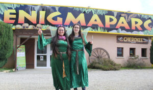 Les Fées d'Enigmaparc devant le parc : deux femmes en robe verte avec des ailes de fées et une baguette magique devant l'entrée d'un parc de loisirs.